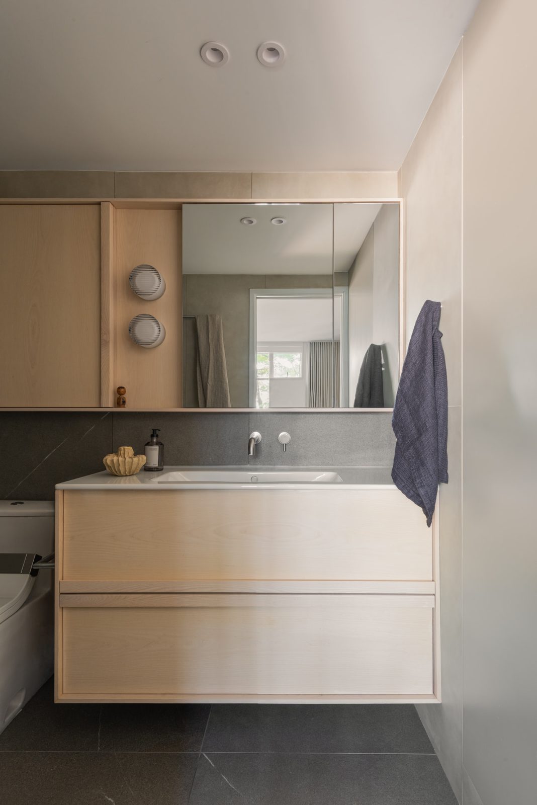 A bathroom with a pale ash vanity, a mirrored cabinet above and dark stone tiling