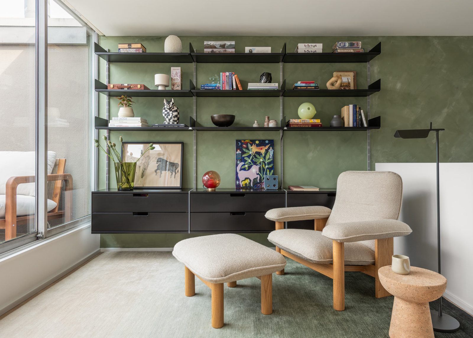 A wall of black shelving and drawers against a green plaster wall, with a lounge chair in front