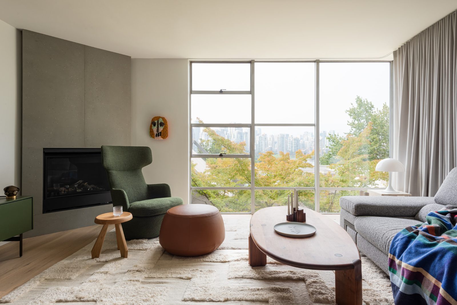 A living room with a poured concrete fireplace wall and full height glazing onto trees and a city skyline